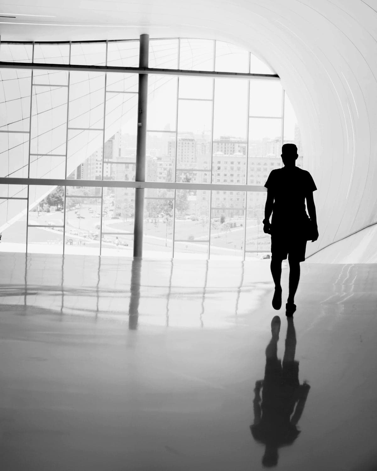 A person in silhouette walks along a curved white corridor towards a wall of windows