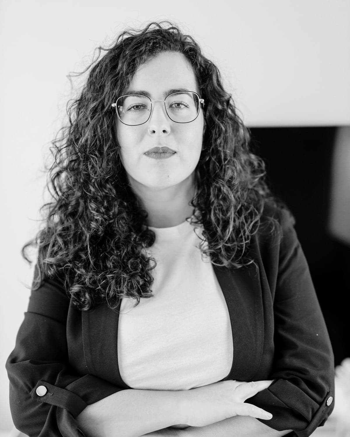 A woman with curly hair and glasses, arms folded, looking straight at the camera