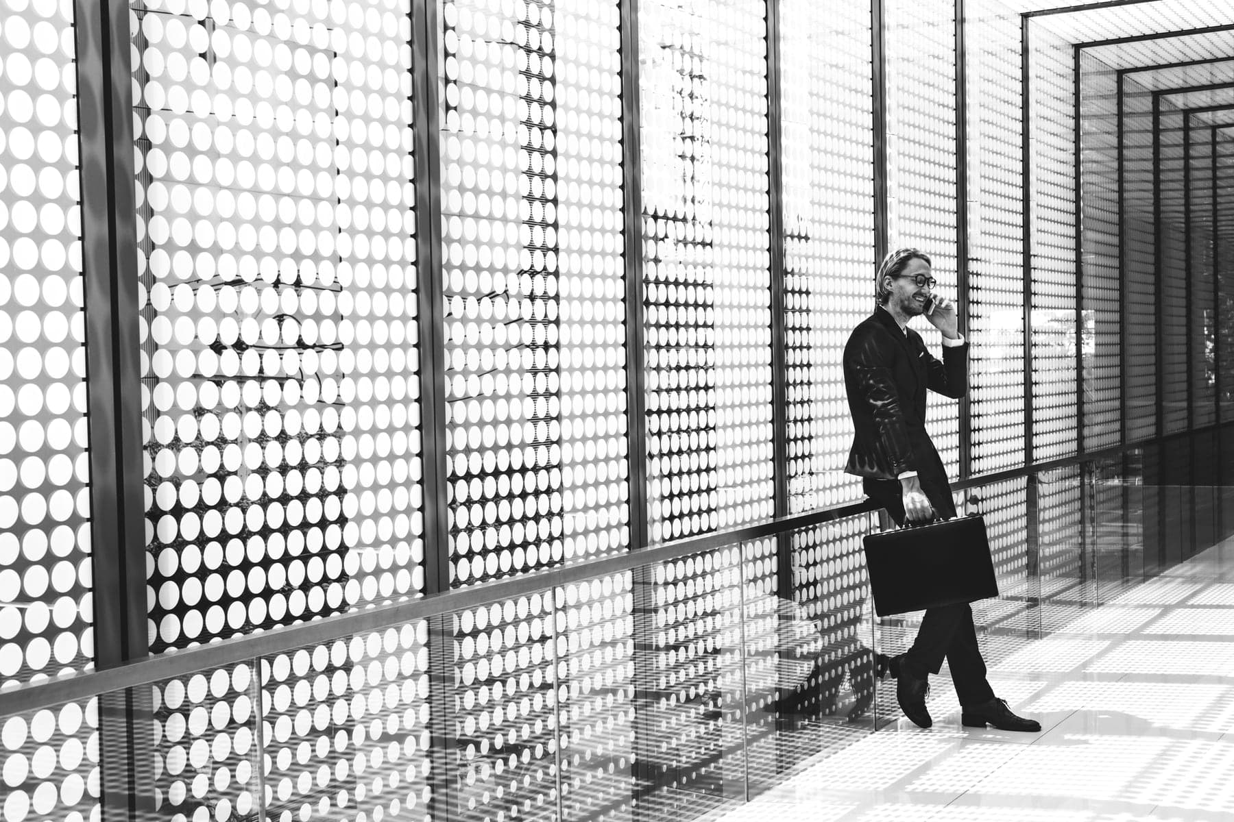 A man in a suit and glasses, briefcase in hand, smiles on a phone call against a wall of round perforations