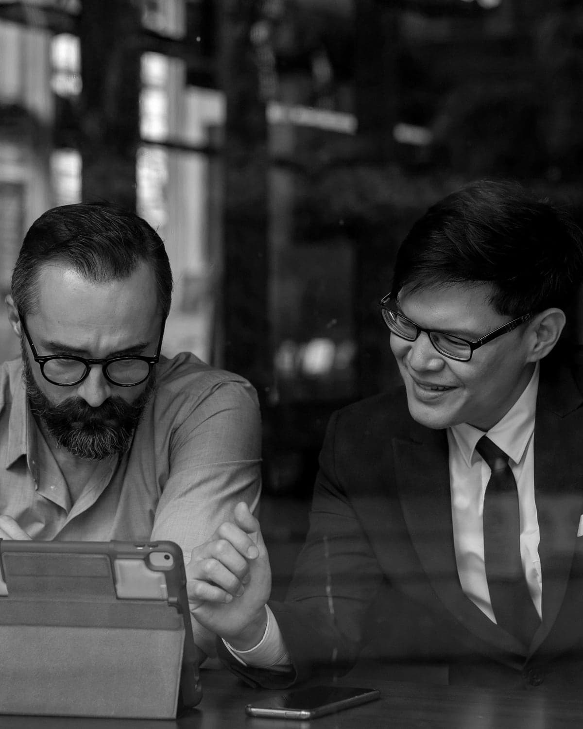 Two men in a café window lean over a tablet, one pointing at the screen as the other smiles
