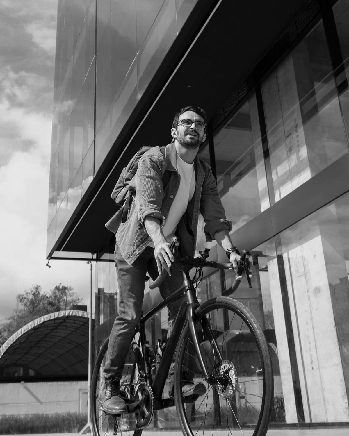A man in glasses rides a bicycle out from under a glass building, looking up