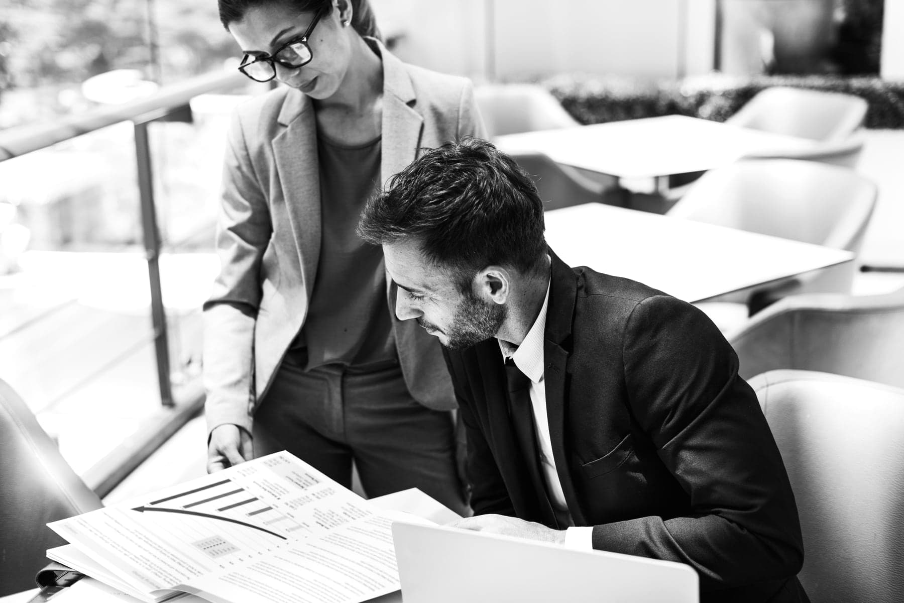 A woman in glasses stands beside a seated colleague as they read through a report with charts