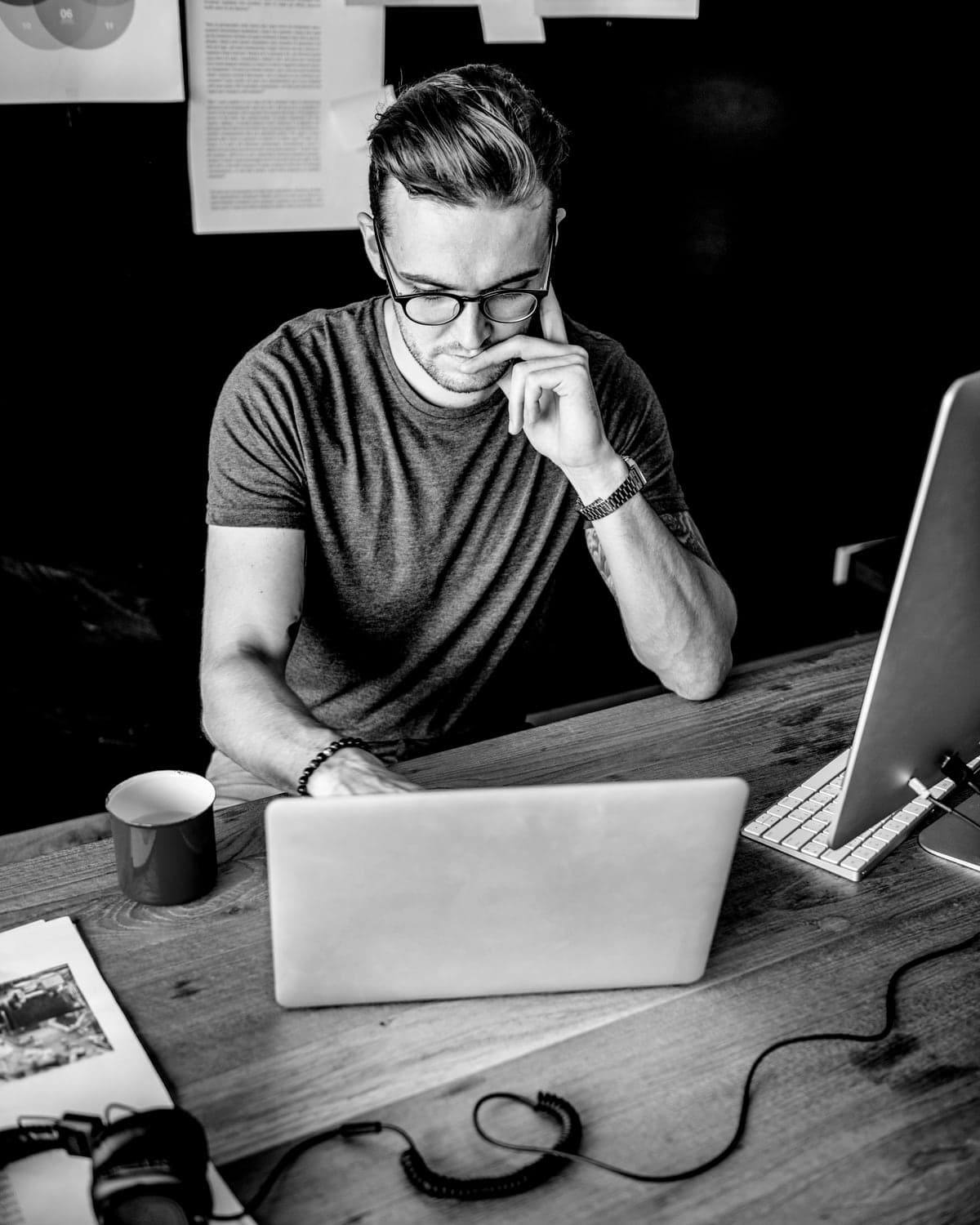 A man in glasses at a laptop, hand to his chin, thinking